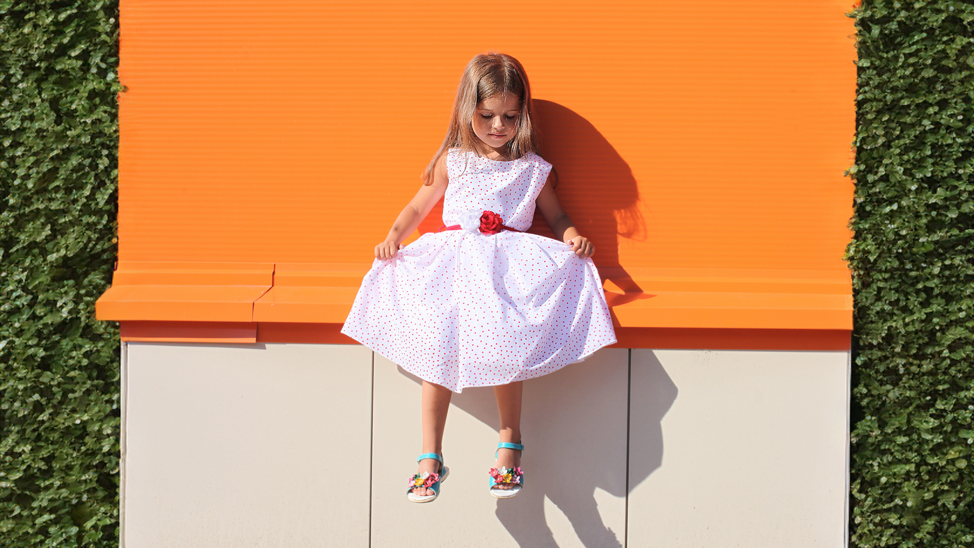 Fille qui porte une robe rose little celebs. L'enfant est assis sur un muret avec un fond orange et des plantes sur les cotés
